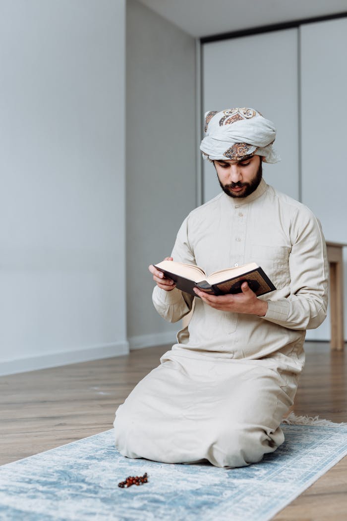 services-bg A man in traditional wear reads the Quran while kneeling on a prayer mat indoors.